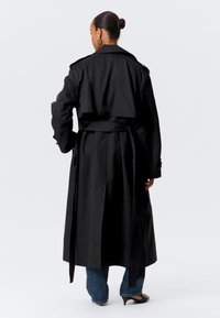 Cappotto trench nero con vita cintata, caratterizzato da un design doppiopetto, ampi rever e uno spacco posteriore. Texture liscia e stile a lunghezza media.
