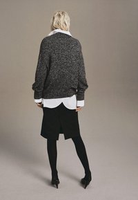 Př oversized šedý pletený svetr, vrstvený přes bílou košili, spárovaný s černou sukní. Obsahuje dlouhé rukávy, žebrované manžety a vysoké podpatky.