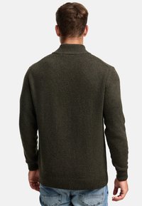 Hombre vestido con un suéter de lana verde oscuro y jeans azules, de pie de espaldas contra un fondo claro y liso.