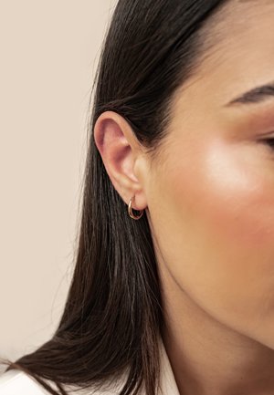 Nahaufnahme eines Frauenohrs mit glattem dunklem Haar und kleinem goldenen Creolen-Ohrring vor neutralem Hintergrund.