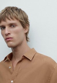 Chemise en lin marron avec un col classique, dotée d'une patte de boutonnage, d'une texture lisse et d'une coupe décontractée, portée par un jeune mannequin masculin.