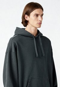 Felpa con cappuccio oversize di colore grigio scuro, dotata di cordino, tasca frontale e dettagli di cucitura visibili. Tessuto morbido con vestibilità comoda.