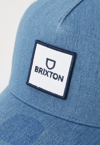 Blue denim cap featuring a white rectangular patch with navy border, displaying "BRIXTON" in bold lettering. Smooth texture, structured design.