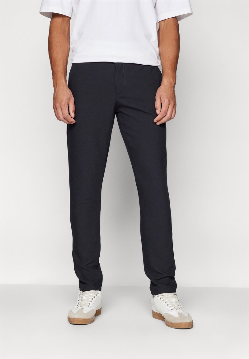 Homme portant un pantalon noir slim, une chemise blanche à manches courtes et des baskets blanches avec des semelles marron clair, debout devant un fond uni.