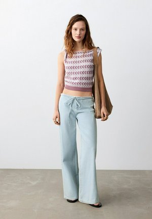 Tânără purtând un top fără mâneci cu model, pantaloni largi de culoare albastru deschis și sandale negre, cu o geantă mare maro atârnându-i pe umăr.