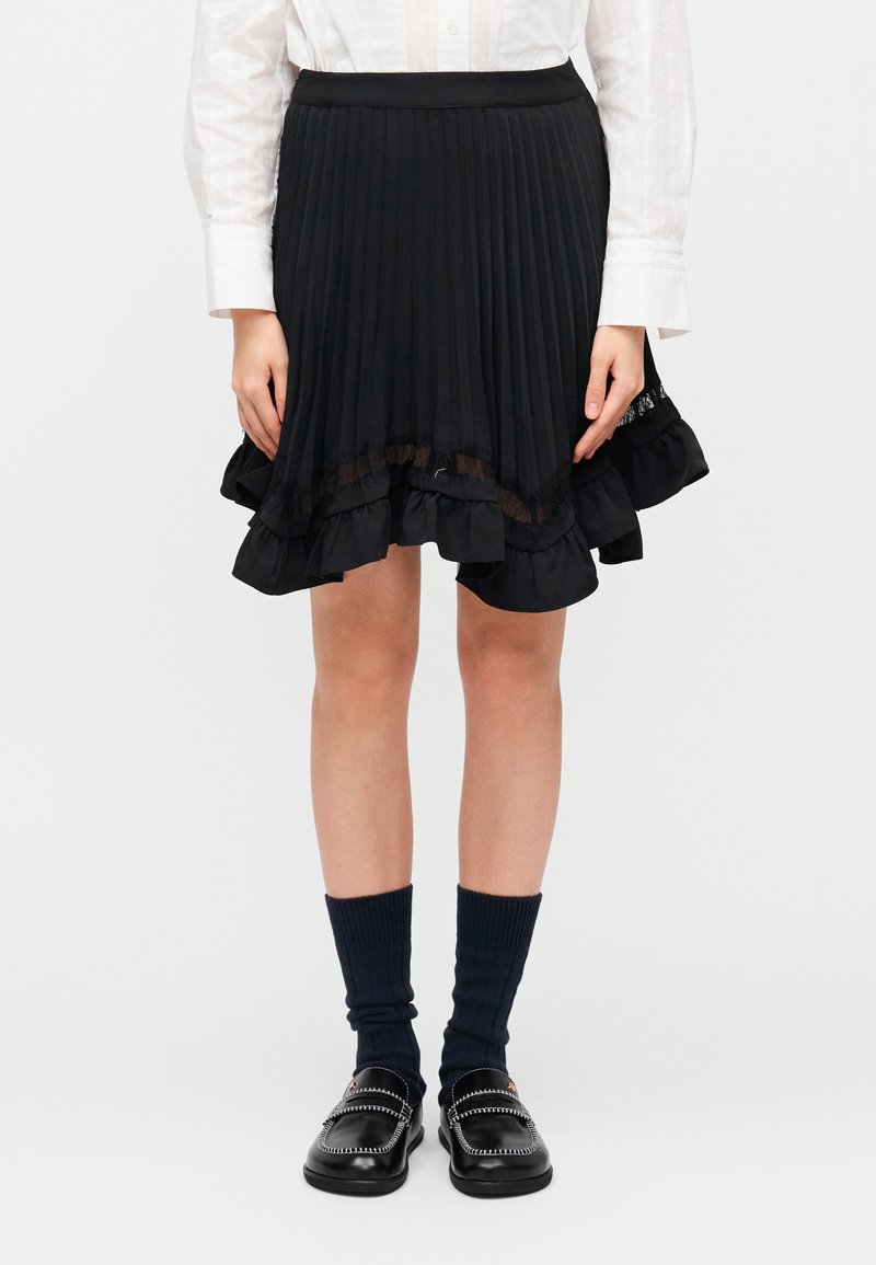 Μαύρη πλισέ φούστα με βολάν δαντέλας στο τελείωμ α, συνδυασμένη με ναυτικομπλε κάλτσες και μαύρα μοκασίνια, με λείο ζωνάκι και στρωματοποιημένο σχέδιο.