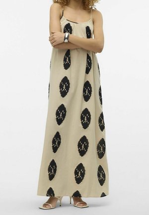 Vrouw draagt een beige maxi-jurk met zwarte abstracte ovale prints, dunne bandjes, zilveren armband en beige sandalen met hakken, staand met gekruiste armen.
