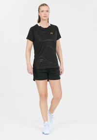 Svart atletisk skjorta med ett subtilt vågmönster, korta ärmar och vit krage; kombinerad med svarta shorts. Bärs med vita sneakers.