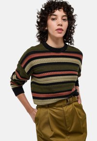 Pull en tricot avec des rayures horizontales en olive, noir, rouille et beige. Doté d'un col rond et de poignets côtelés, porté avec un pantalon taille haute.