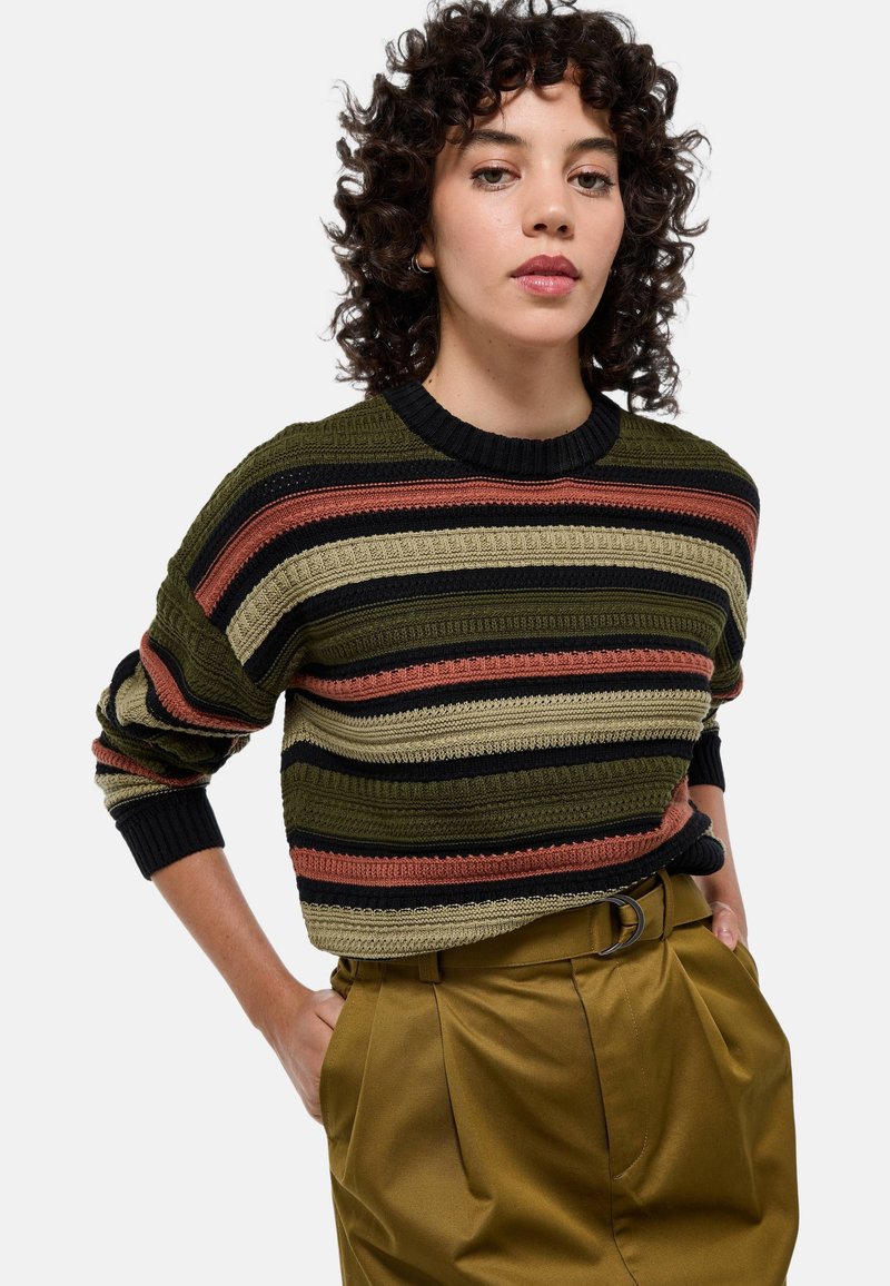 Pull en tricot avec des rayures horizontales en olive, noir, rouille et beige. Doté d'un col rond et de poignets côtelés, porté avec un pantalon taille haute.