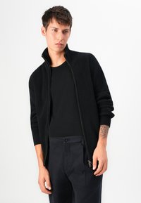 Giovane uomo con capelli corti, che indossa un maglione nero a coste con zip, una t-shirt nera e pantaloni scuri, che mostra un tatuaggio sul suo avambraccio sinistro.