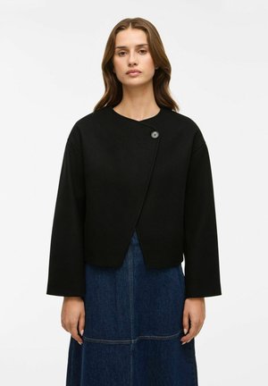 Veste noire courte avec un col rond, fermeture à bouton unique à l'avant et manches larges, associée à une jupe en denim bleu.