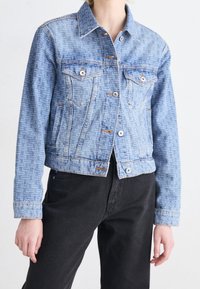 Primer plano de una persona que lleva una chaqueta vaquera azul claro con estampado, botones plateados y pantalones negros, de pie frente a un fondo liso.