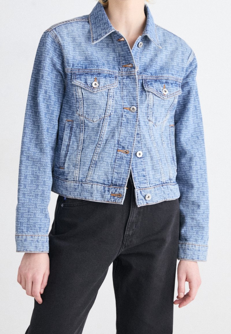 Närbild på en person som bär en ljusblå mönstrad denimjacka med silverknappar och svarta byxor, stående mot en enfärgad bakgrund.