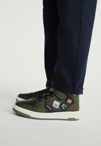Sneakers verdi e nere con finitura strutturata, caratterizzate da toppe, una chiusura in Velcro e una suola intermedia bianca screziata. Design casual con pantaloni a risvolto.