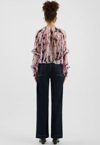 Losse roze blouse met een tie-dye patroon en ruches aan de mouwen, gecombineerd met donkergroene wijduitlopende spijkerbroek en zwarte laarzen met hak.