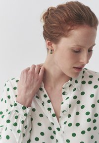 Blusa bianca a pois verdi, con un design a bottoni e colletto. La modella indossa orecchini a perno colorati e geometrici.