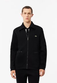 Chaqueta negra con cremallera hecha de una tela texturizada, con un cuello, dos bolsillos laterales y un pequeño logo verde en la parte izquierda del pecho.