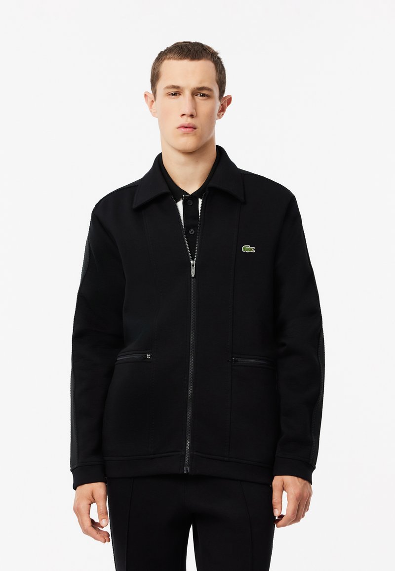 Chaqueta negra con cremallera hecha de una tela texturizada, con un cuello, dos bolsillos laterales y un pequeño logo verde en la parte izquierda del pecho.