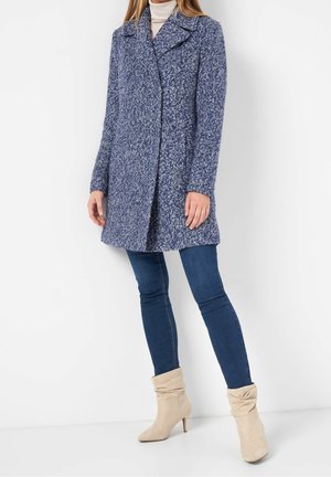 Kobieta ubrana w niebieski teksturowany płaszcz do kolan, ciemnoniebieskie obcisłe jeansy, beżowe luźne botki na obcasie oraz biały golf.