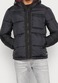 Pánská černá prošívaná bunda s kapucí, s prošitou texturou, předním zipem, kapsou na hrudi a černým lemováním podél zipu.