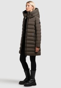 Brązowa ocieplana parka z wysokim kołnierzem, zamkiem błyskawicznym i bocznymi kieszeniami. Tkanina o fakturze i długie rękawy. W zestawie z czarnymi botkami za kostkę.