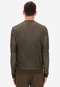 Uomo con capelli corti e scuri che indossa un maglione aderente a coste verde oliva e pantaloni abbinati, visto di spalle su uno sfondo neutro.