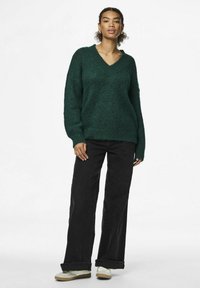 Pārmērīgas, dziļi zaļas adītas džemperis ar V veida kaklu un ribotām piedurknēm; saskaņots ar augsto vidukli melnām platām biksēm un baltām sporta kedām.