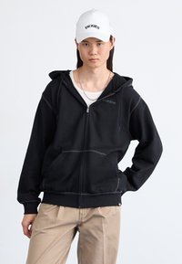 Crna zip up dukserica sivo šavovima, prednjim džepovima i kapuljačom. Uparena sa bež hlačama. Bijela kapica s imenom branda "DICKIES."
