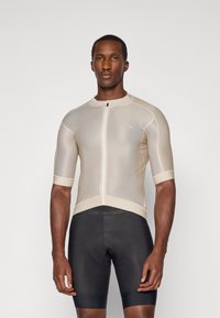 Männlicher Model trägt ein beiges kurzärmliges Radtrikot mit Reißverschluss vorne und schwarze gepolsterte Radhosen, steht vor einem schlichten hellen Hintergrund.