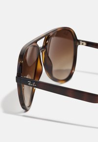 Occhiali da sole tartaruga marrone con lenti sfumate, con il logo nero Ray-Ban sulla tempia. Montatura in plastica liscia, lenti rotonde.
