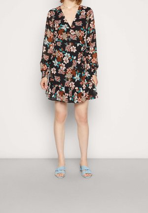 Femme portant une robe portefeuille noire à fleurs, manches longues et ourlet volanté, associée à des sandales à talons ouvertes bleu clair.