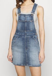 Kvinna som bär en urblekt blå denimoverallklänning med ficka fram och silverknappdetaljer över en ljusgrå ärmlös topp.