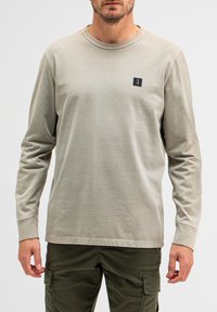 Helles beige Langarm-T-Shirt aus Baumwollmischung mit Rundhalsausschnitt. Verfügt über ein kleines schwarzes Logopatch auf der linken Brust. Einfaches Design.