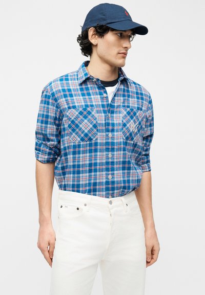 Νεαρός άνδρας που φοράει μπλε πουκάμισο με καρό κουμπιά, λευκό παντελόνι και μπλε σκούφο μπέιζμπολ, κοιτάζοντας προς τα δεξιά του.