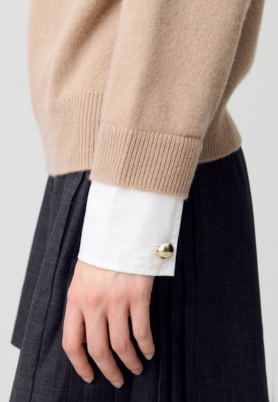 Pull en cachemire beige avec des poignets côtelés, porté sur une chemise blanche à boutons avec un bouton en or. Jupe plissée sombre visible en dessous.