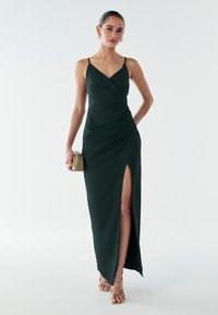 Smaragdgrünes Satinkleid für den Abend mit Wickeldesign, seitlichem Schlitz und dünnen Trägern. Das Modell hält eine hellgoldene Clutch und trägt filigrane Sandalen.