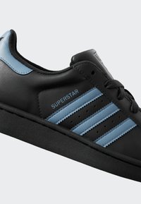 Black sneaker with light blue stripes and heel tab, "SUPERSTAR" text on side, black laces, and textured black sole.
