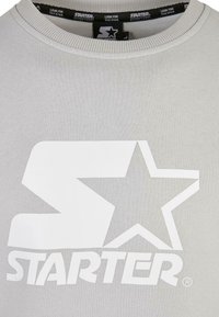 Sweat-shirt gris avec un grand logo "Starter" blanc comportant un "S" stylisé et une étoile sur la poitrine, ainsi qu'un col intérieur de marque.
