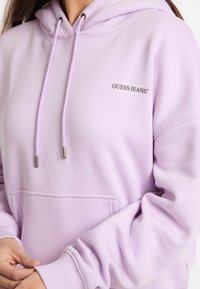 Felpa lavanda realizzata in morbido tessuto, con tasca a canguro, cordini con estremità metalliche e un piccolo logo ricamato "GUESS JEANS".