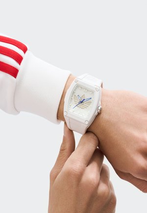 Persona pielāgo baltu Adidas rokas pulksteni ar ziliem rādītājiem, valkājot baltu jaku ar sarkaniem svītrām uz piedurknēm.