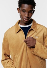 Giacca in suede beige con chiusura a zip e colletto, caratterizzata da un dettaglio ricamato "NYC" sul petto e una texture morbida.