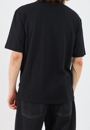 Person trägt ein schlichtes schwarzes kurzärmeliges T-Shirt und dunkle Jeans, von hinten vor weißem Hintergrund gezeigt.