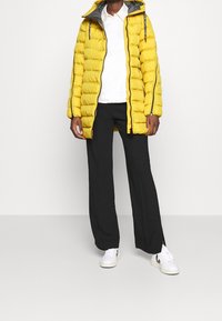 Abrigo puffer amarillo con capucha y cremallera lateral, combinado con pantalones negros de pierna ancha y zapatillas blancas con detalles negros.