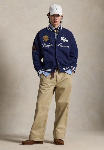 Ναυτικό varsity μπουφάν με λευκές ρίγες, κέντημα "Ralph Lauren" και λογότυπο λιονταριού· ανοιχτό μπλε πουκάμισο, χακί παντελόνι και καφέ παπούτσια.