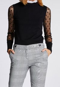 Schwarzer Pullover mit durchsichtigen gepunkteten Ärmeln, kombiniert mit grauen Karo-Hosen, die Taschen und Knopfakzente aufweisen.