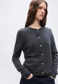 Γυναίκα με μακριά καστανά μαλλιά, που φοράει σκούρα γκρι ζακέτα με κουμπιά και σκούρα τζιν, τα χέρια στις τσέπες, κοιτάζοντας ελαφρώς προς το πλάι.