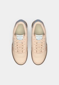 Sneakers beige chiaro con una texture liscia, caratterizzati da accenti blu sulla linguetta, design con lacci e suola in gomma marrone chiaro.