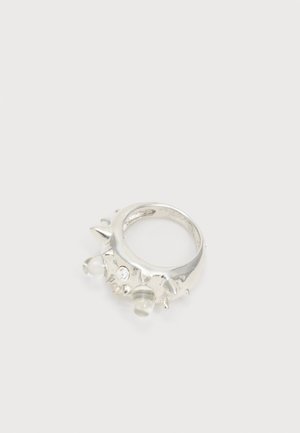 Inel de argint cu mai multe țepi, cristal mic și transparent și o decorațiune rotunjită transparentă pe un fundal simplu.