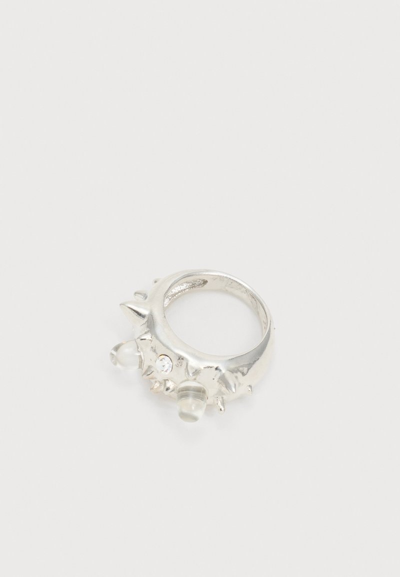 Ασημένιο δαχτυλίδι με πολλαπλές αιχμές, μικρό διαυγές κρύσταλλο και στρογγυλεμένο διαφανές στολίδι σε μονόχρωμο φόντο.